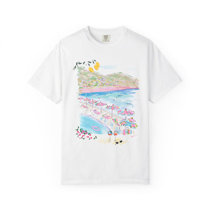 White t-shirt featuring a colorful hand-drawn Amalfi Coast beach club scene with striped umbrellas, blue Mediterranean water, and hillside villages, illustrated by Rebecca Illustrated.