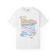 White t-shirt featuring a colorful hand-drawn Amalfi Coast beach club scene with striped umbrellas, blue Mediterranean water, and hillside villages, illustrated by Rebecca Illustrated.