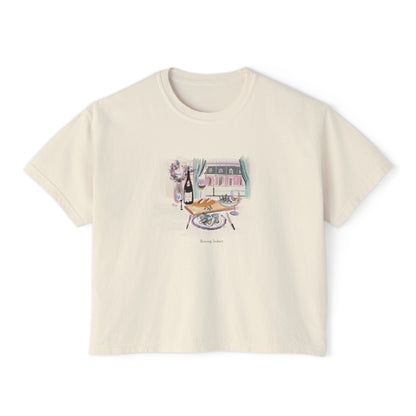 Cream cropped t-shirt featuring a hand-painted brunch scene with oysters, champagne, flowers, and a Parisian-style window view, with the text ‘Morning Indulge,’ illustrated by Rebecca Illustrated."