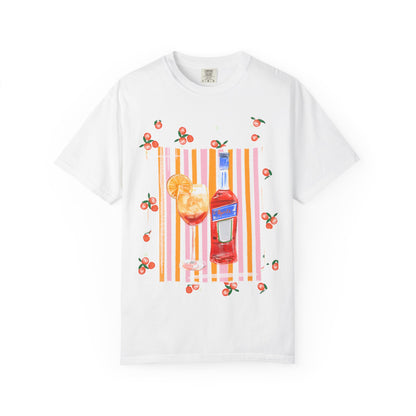 White t-shirt with a hand-painted Aperol spritz cocktail illustration, orange slice garnish, striped background, and scattered citrus details, designed by Rebecca Illustrated.