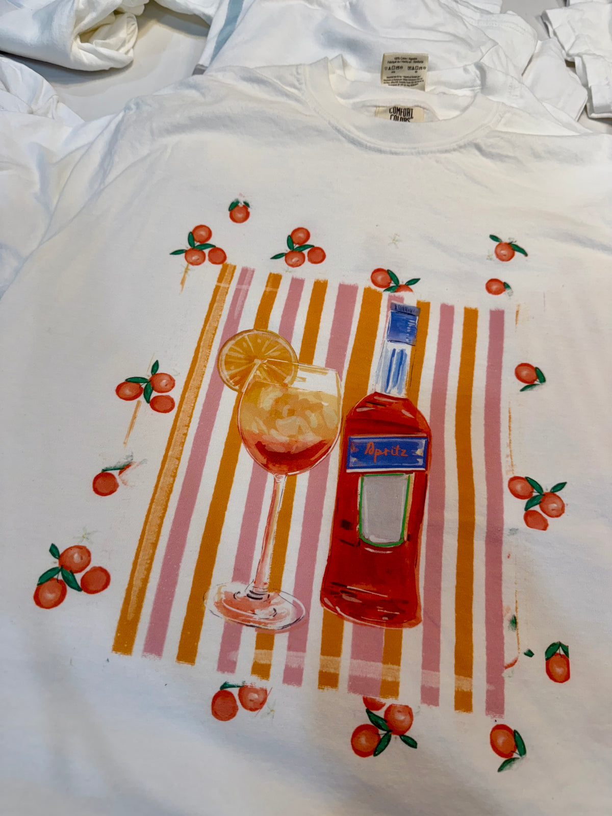 White t-shirt with a hand-painted Aperol spritz cocktail illustration, orange slice garnish, striped background, and scattered citrus details, designed by Rebecca Illustrated.