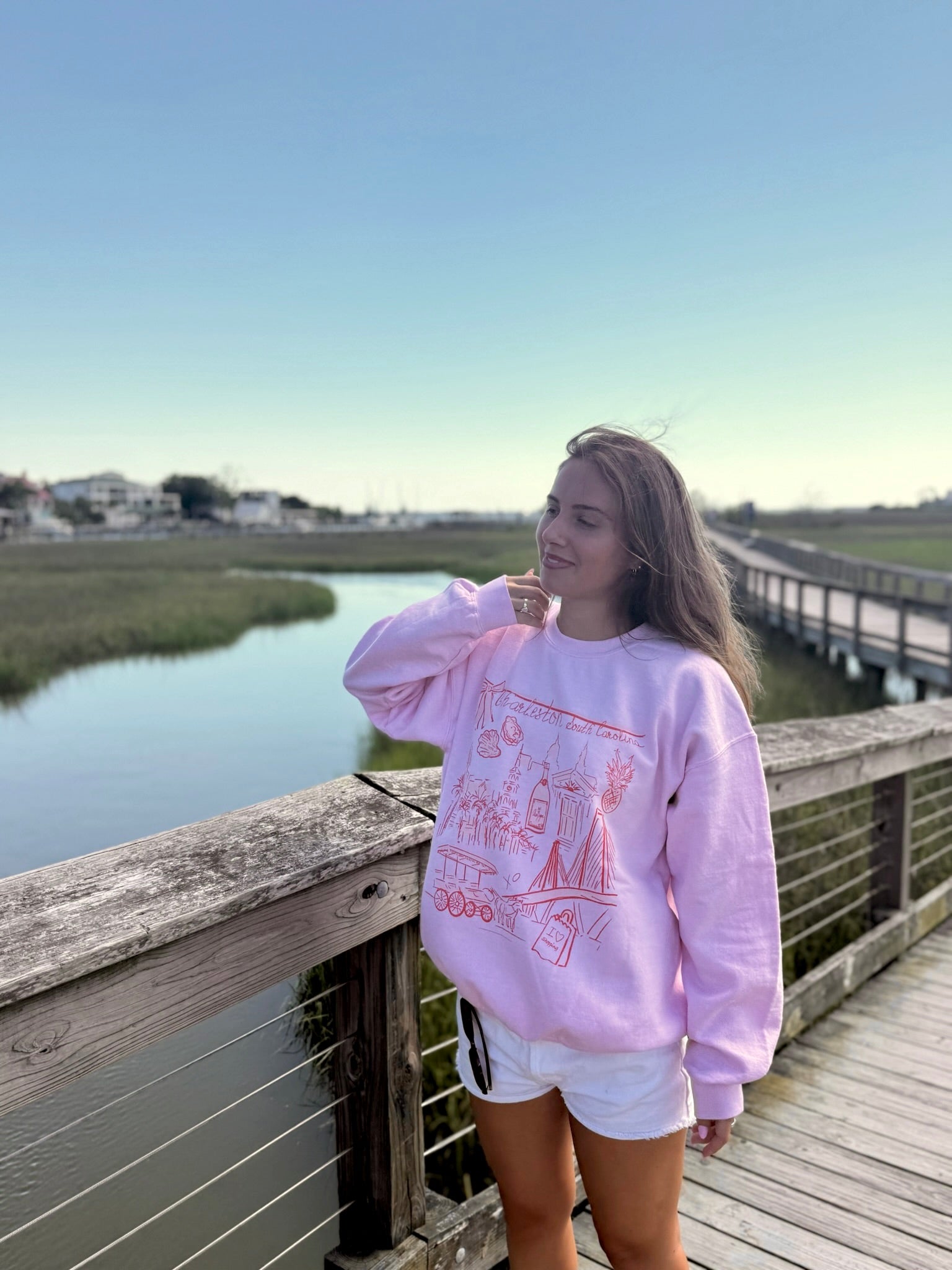 Person wearing a pink sweatshirt with text and graphics, standing on a wooden bridge by a body of water.
