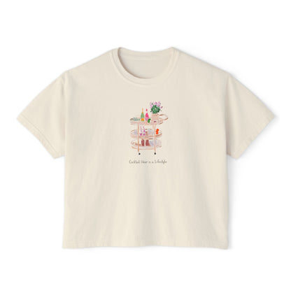 Cream cropped t-shirt featuring a hand-painted bar cart with cocktails, flowers, glassware, and the text ‘Cocktail Hour is a Lifestyle,’ illustrated by Rebecca Illustrated.