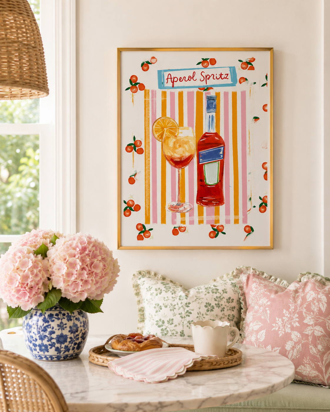 A framed art print featuring a colorful illustration of an Aperol Spritz cocktail and bottle, displayed on a cream wall in a bright, styled room with pink flowers, pillows, and a marble table.