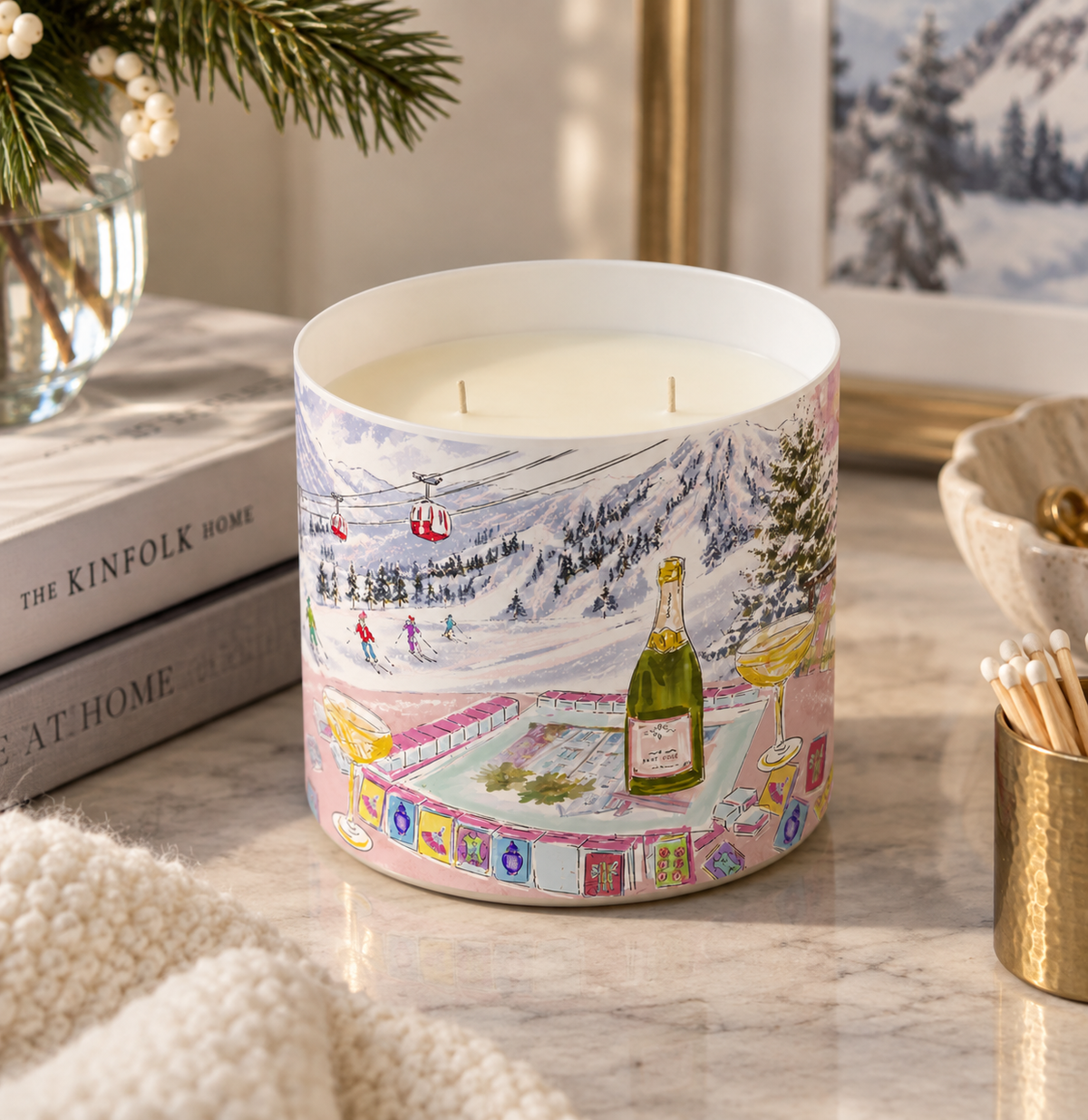 A cylindrical scented candle with a winter ski resort illustration sits on a marble surface in a styled home setting. The candle features a colorful print of snowy mountains, ski lifts, a ski slope, and a champagne bottle, with white wax visible inside.