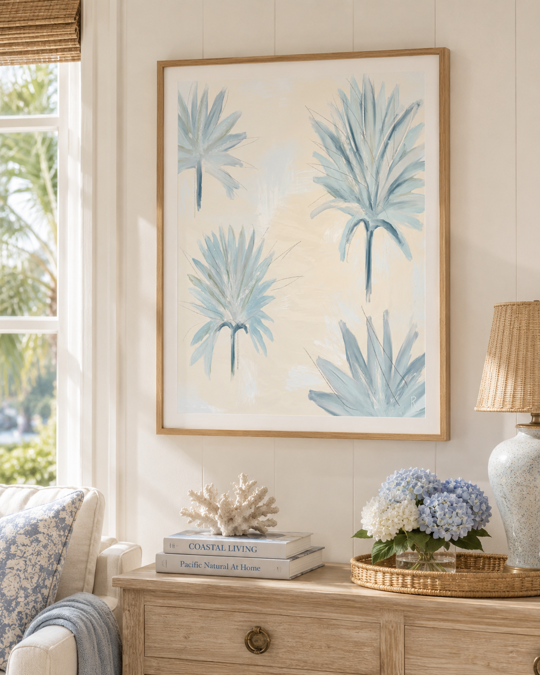 A framed botanical art print featuring light blue palm fronds on a cream background, styled in a bright, coastal-themed living room above a wooden console table.