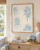 A framed botanical art print featuring light blue palm fronds on a cream background, styled in a bright, coastal-themed living room above a wooden console table.