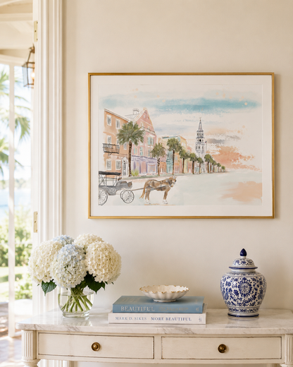A framed watercolor art print depicting a coastal town street scene with a horse-drawn carriage, hung on a cream wall above a console table with flowers, books, and a blue-and-white ceramic jar.