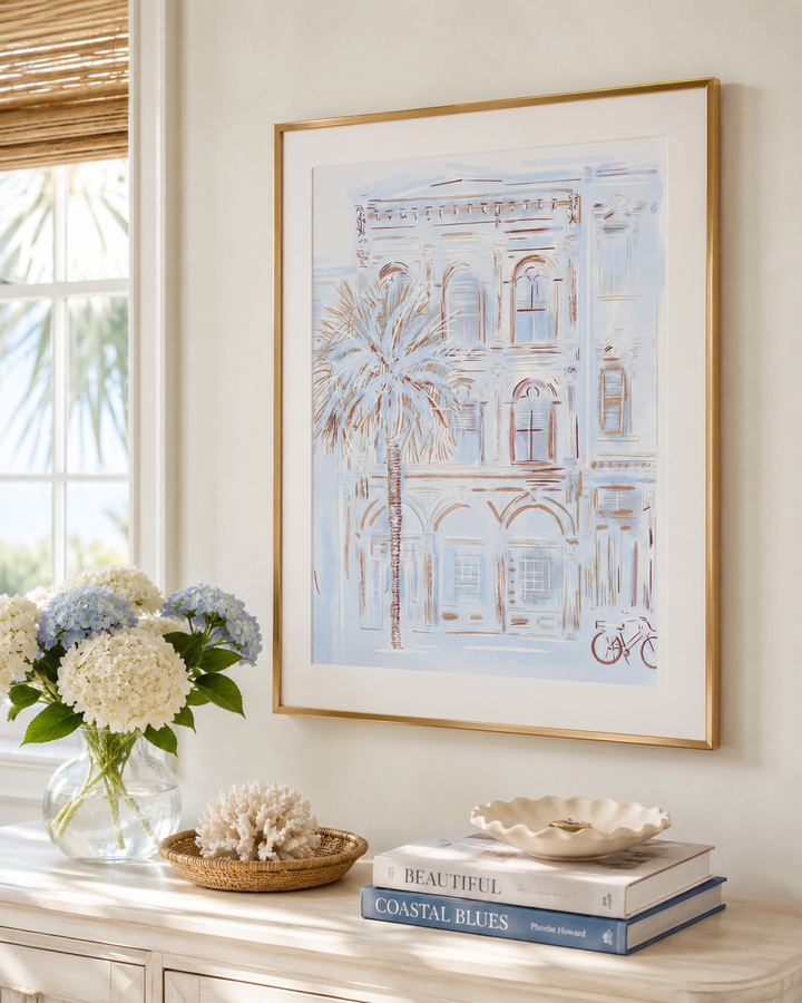 A framed art print featuring a light blue and white architectural illustration with a palm tree, displayed on a console table in a bright room next to a window with a woven shade. The print is styled with a vase of hydrangeas, a woven basket, and books.