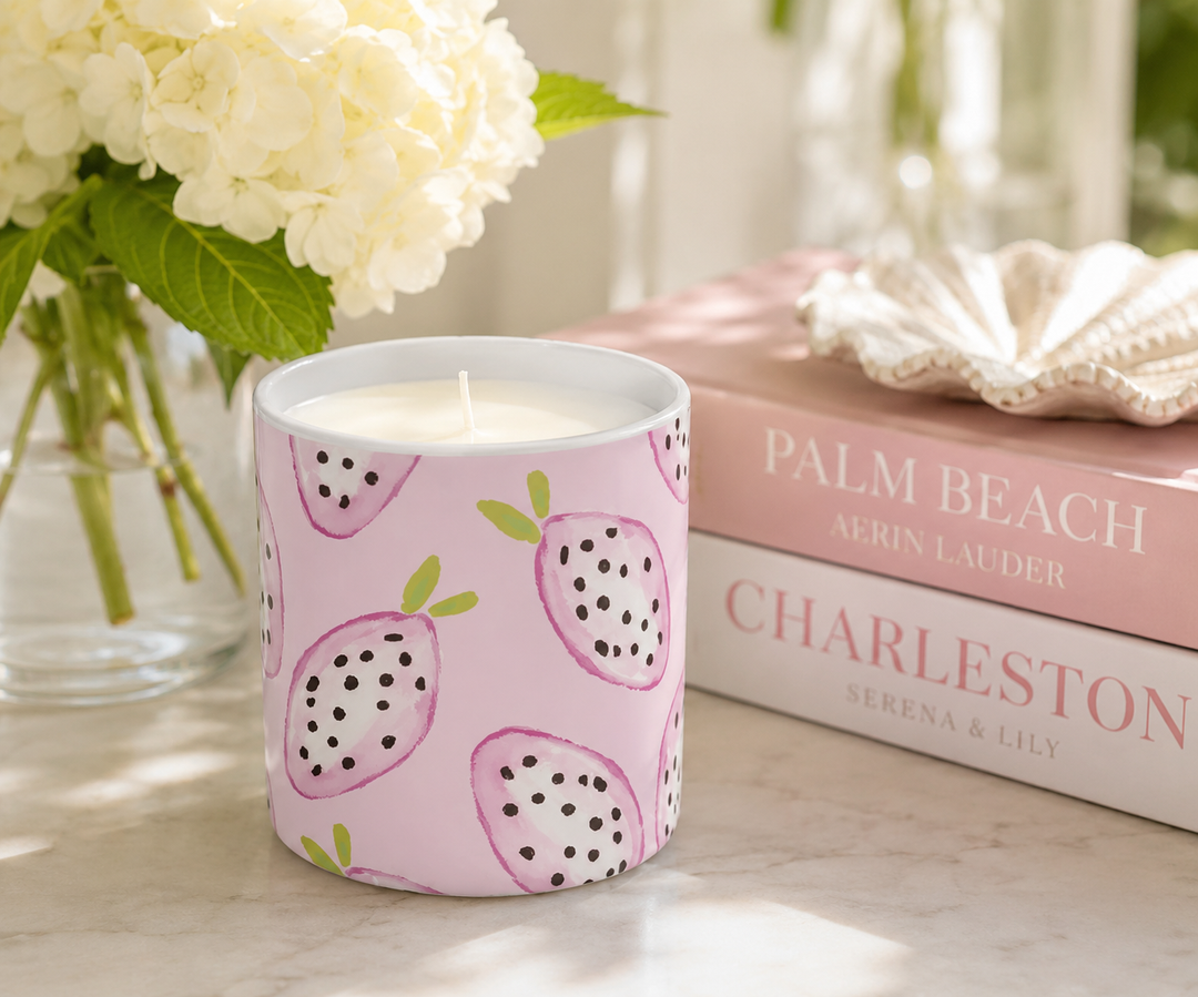 A pink scented candle with a dragon fruit pattern sits on a marble surface next to a vase of white flowers and a stack of books.