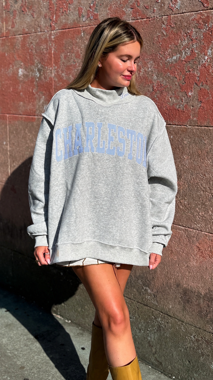 A woman wearing an oversized light gray sweatshirt with 'CHARLESTON' printed in light blue arched letters across the chest, standing against a textured reddish-brown wall.