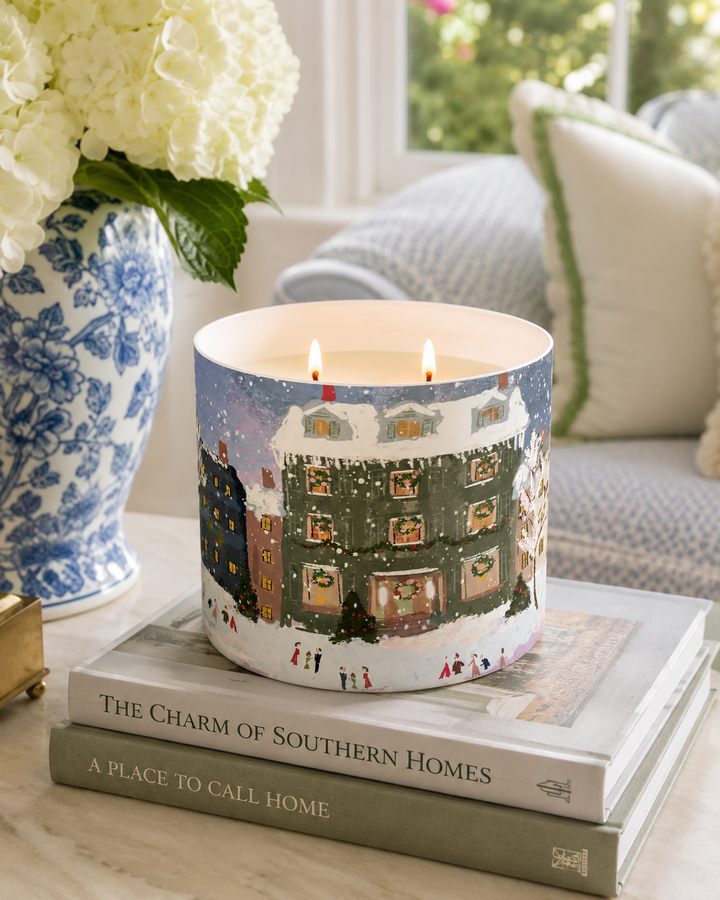 A lit double-wick candle in a cylindrical holder printed with a winter village scene, styled on stacked books in a bright living room setting.