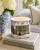A lit double-wick candle in a cylindrical holder printed with a winter village scene, styled on stacked books in a bright living room setting.