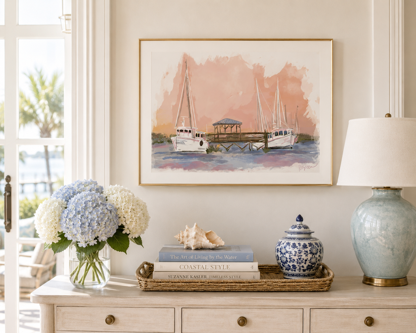 A framed watercolor painting of sailboats at a harbor hangs on a cream wall above a console table styled with a vase of hydrangeas, books, a shell, a blue-and-white lidded jar, and a table lamp.