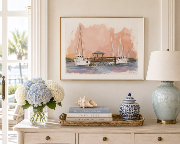 A framed watercolor painting of sailboats at a harbor hangs on a cream wall above a console table styled with a vase of hydrangeas, books, a shell, a blue-and-white lidded jar, and a table lamp.