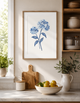 Kitchen interior with a framed blue flower print on the wall, cutting board, and bowl of fruit.