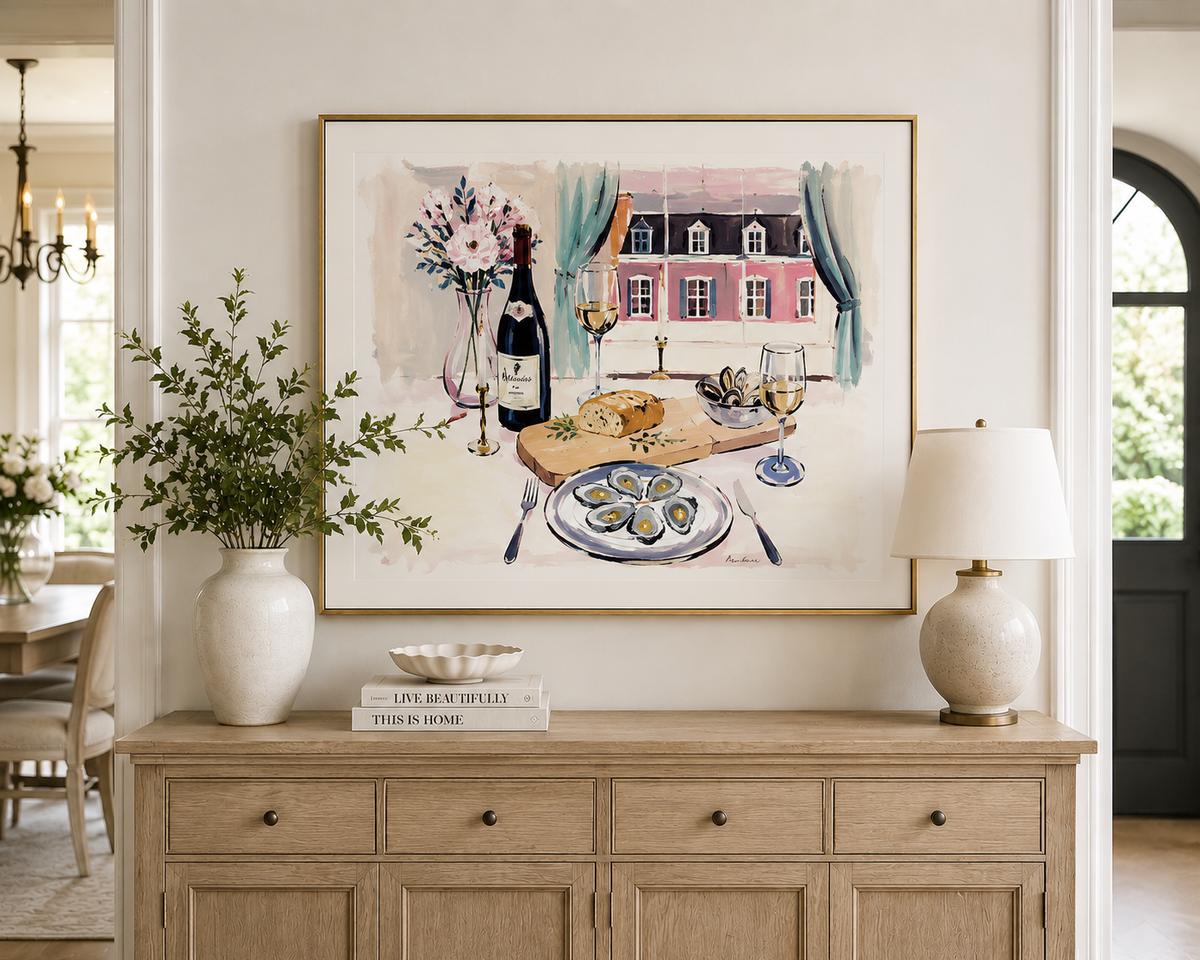 Watercolor-style painting by Rebecca Illustrated featuring oysters, a bottle of wine, and glasses of white wine on a dining table overlooking a pink French château through open blue curtains. Framed artwork displayed above a wooden sideboard in an elegant dining room.