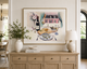 Watercolor-style painting by Rebecca Illustrated featuring oysters, a bottle of wine, and glasses of white wine on a dining table overlooking a pink French château through open blue curtains. Framed artwork displayed above a wooden sideboard in an elegant dining room.