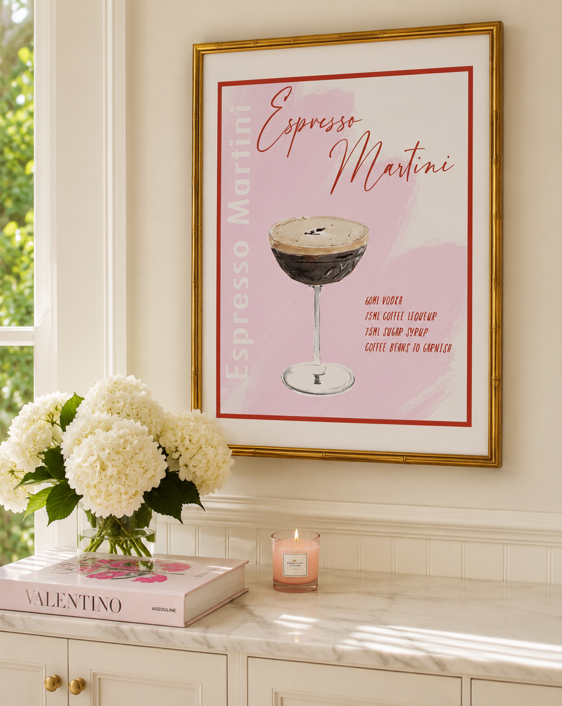 A framed art print featuring an illustration of an Espresso Martini cocktail on a light pink background, hanging on a cream wall above a marble counter with a vase of white flowers and a candle.