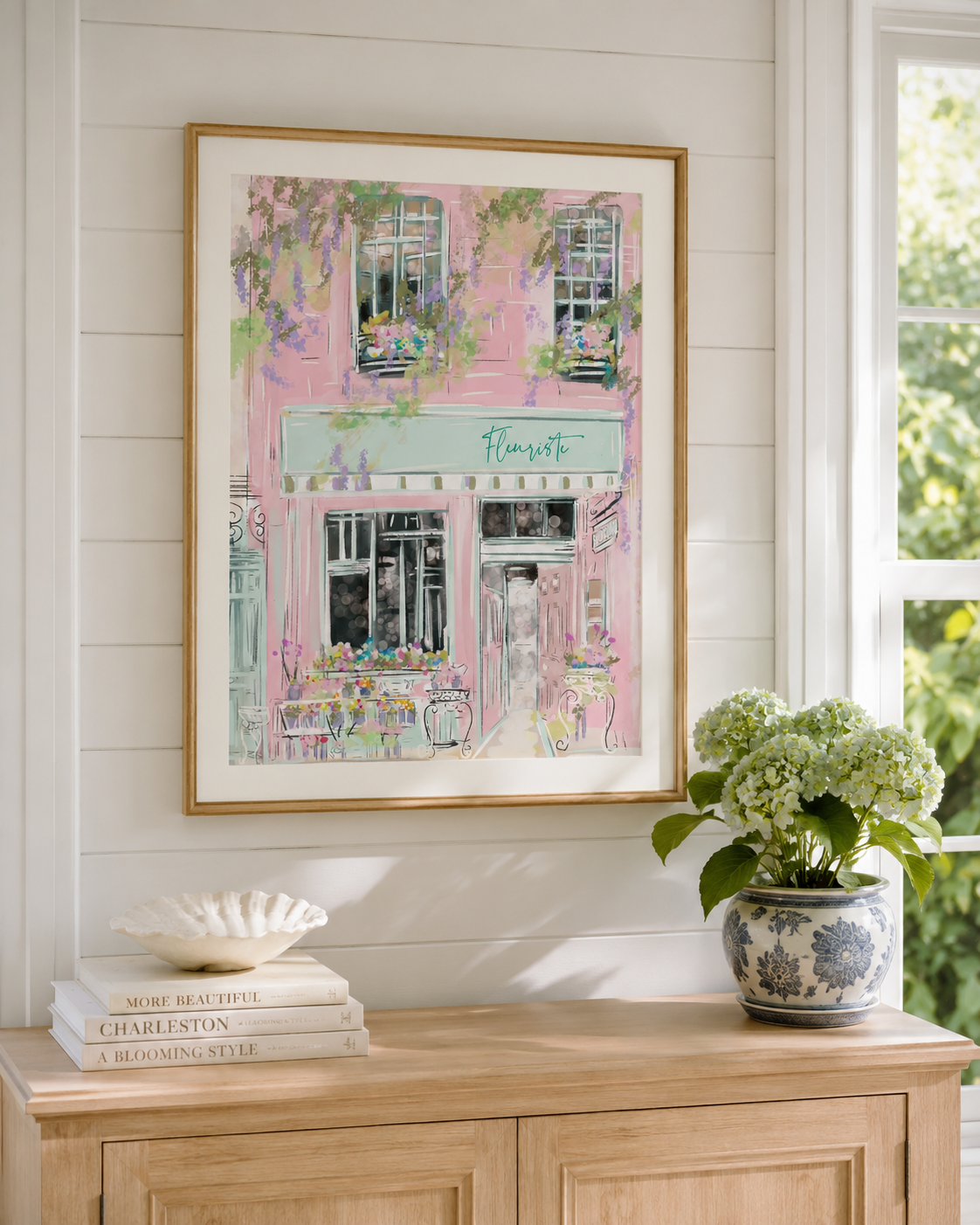 A framed pastel art print of a pink cafe building hangs on a white shiplap wall above a light wood console table styled with books, a shell dish, and a potted hydrangea.