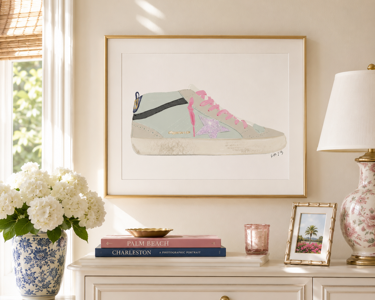 A framed art print featuring a pastel watercolor illustration of a high-top sneaker, displayed on a cream-colored wall above a console table decorated with a blue and white floral vase of white flowers, books, a pink candle, and a floral table lamp.