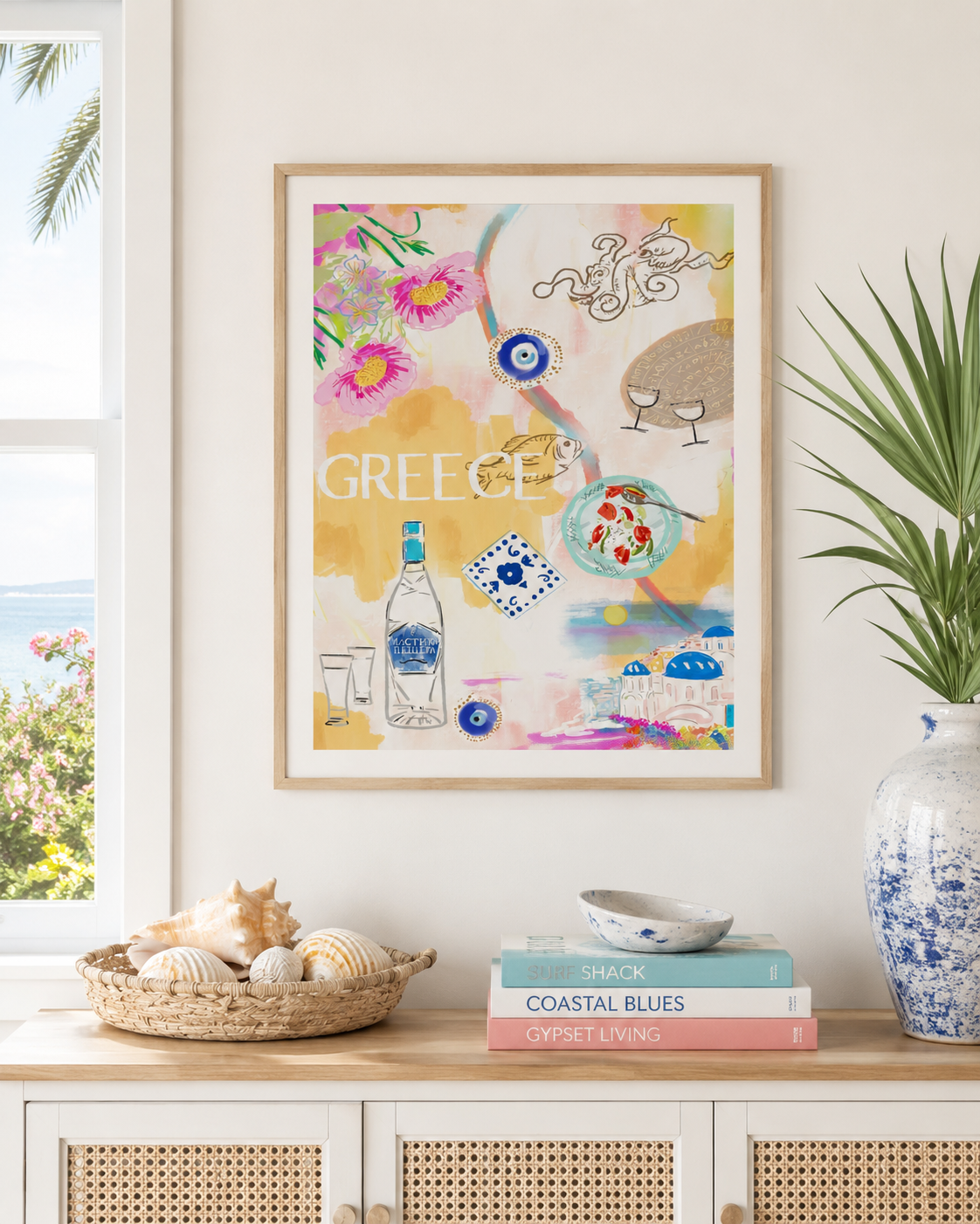 A framed art print featuring a colorful Mediterranean-themed illustration with the word 'GREECE' and coastal motifs, displayed on a white wall above a console table with books, shells, and a potted plant.