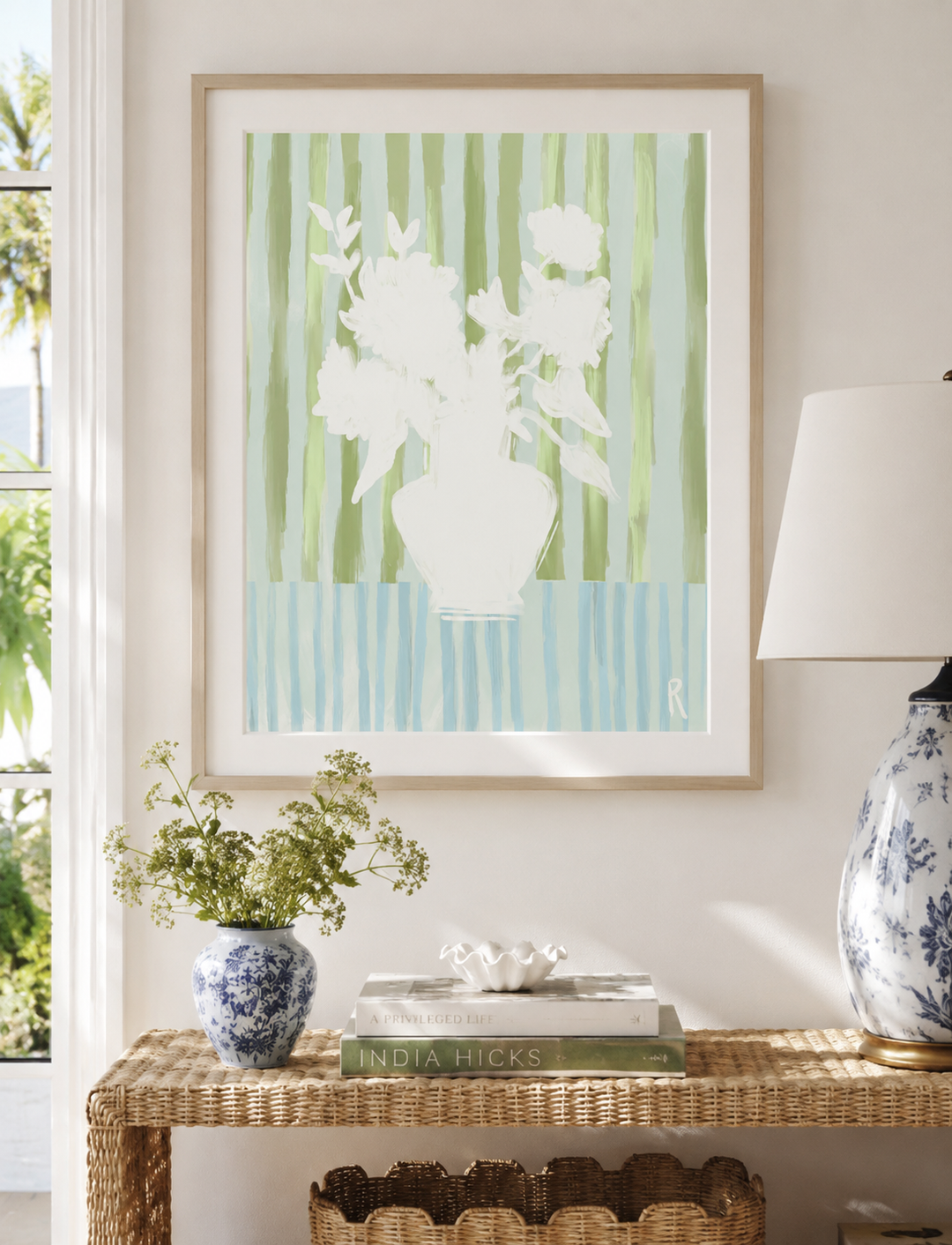 A framed art print featuring a white floral silhouette against vertical green and light blue stripes, displayed on a beige wall above a wicker console table with a lamp, books, and a small vase.