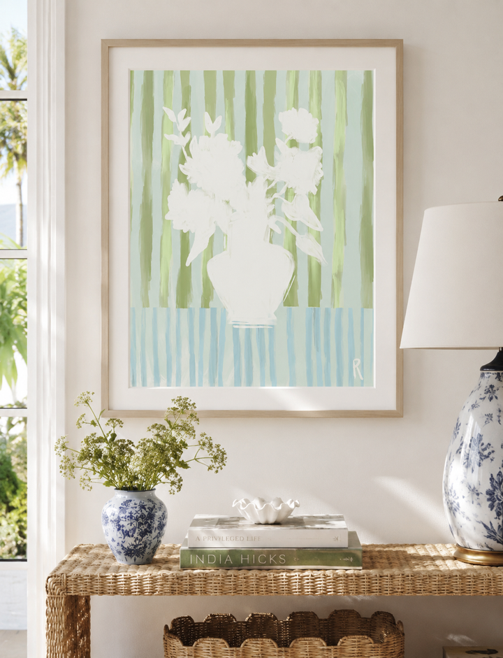 A framed art print featuring a white floral silhouette against vertical green and light blue stripes, displayed on a beige wall above a wicker console table with a lamp, books, and a small vase.