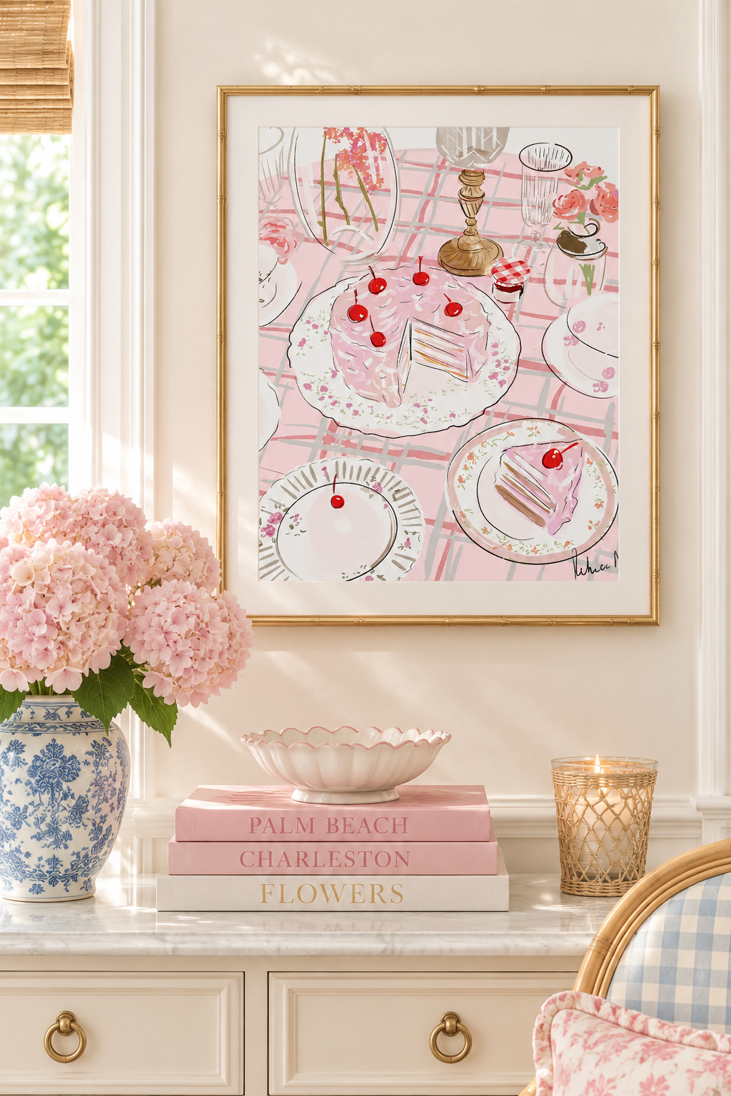 A framed art print featuring a pink and white illustration of a tea and cake table, styled on a cream console table with pink flowers, books, and a candle.