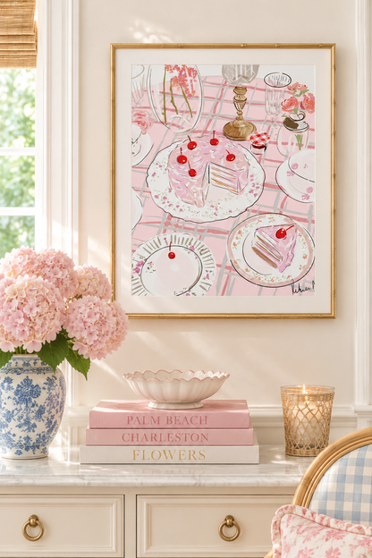 A framed art print featuring a pink and white illustration of a tea and cake table, styled on a cream console table with pink flowers, books, and a candle.