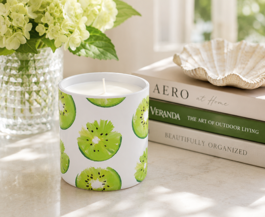 A white ceramic candle jar printed with green kiwi slice patterns, placed on a marble surface next to a vase of pale green hydrangeas and a stack of books.
