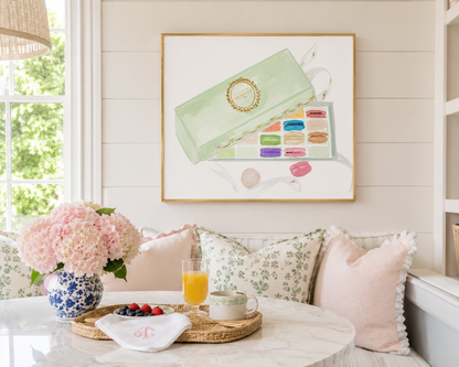 A framed art print featuring a pastel illustration of a green makeup palette and macarons, displayed on a white paneled wall above a cozy seating area with pink pillows and a marble table.