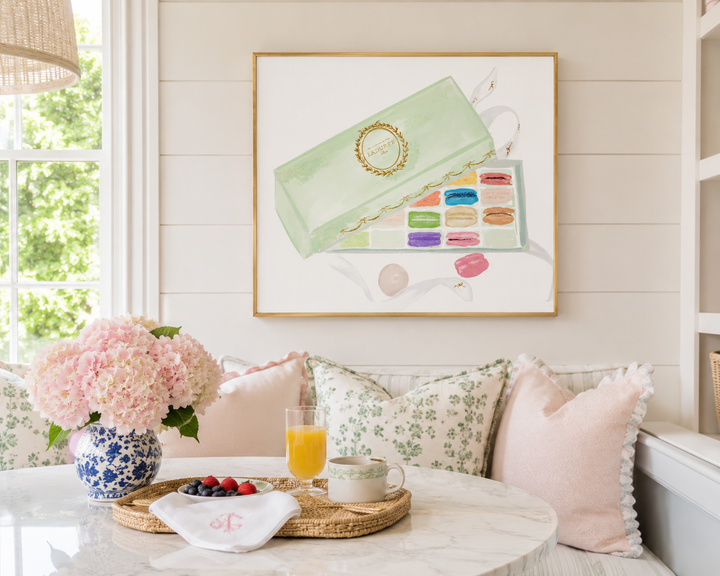 A framed art print featuring a pastel illustration of a green makeup palette and macarons, displayed on a white paneled wall above a cozy seating area with pink pillows and a marble table.