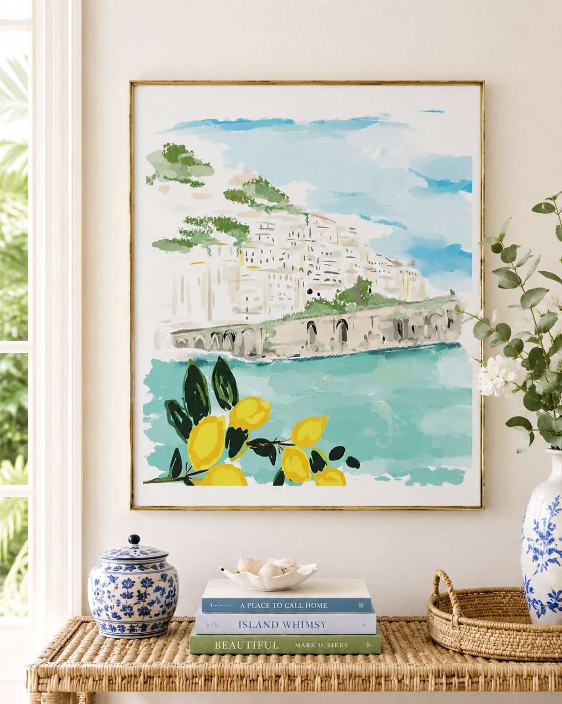 A framed watercolor art print depicting a coastal hillside town with turquoise water and yellow lemons, styled on a woven table in a bright room with books, a blue-and-white ceramic jar, and a vase.