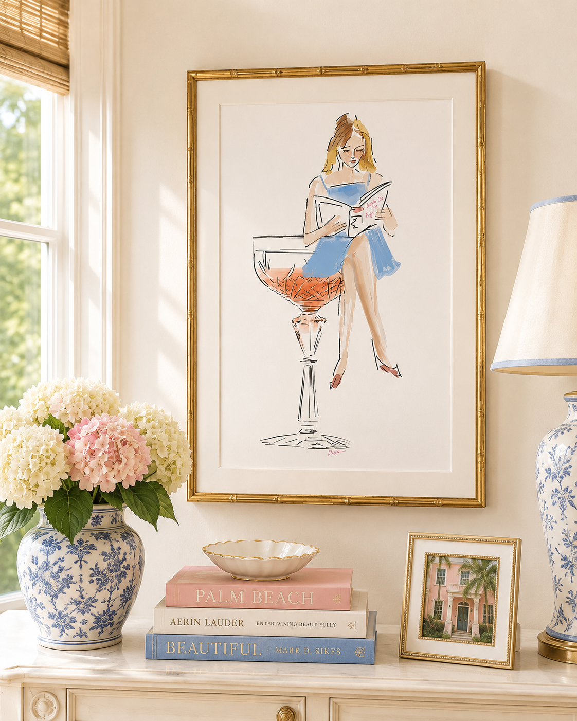 A framed art print featuring a vintagestyle illustration of a woman in a blue dress sitting in a cocktail glass, displayed on a console table with books, a blue and white vase of flowers, and a lamp in a bright room.
