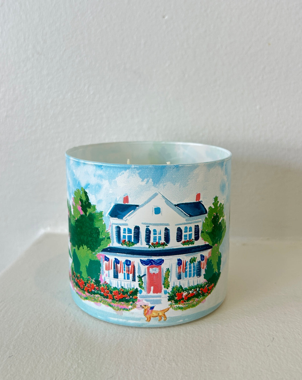 Rebecca Illustrated candle featuring a hand-painted colonial home decorated with American flags, colorful flower gardens, and patriotic Americana details on a frosted glass vessel.
