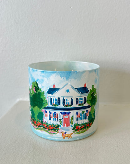 Rebecca Illustrated candle featuring a hand-painted colonial home decorated with American flags, colorful flower gardens, and patriotic Americana details on a frosted glass vessel.