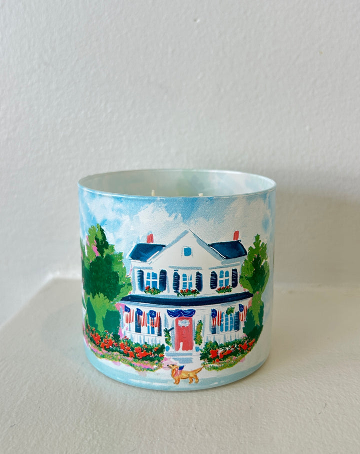 Rebecca Illustrated candle featuring a hand-painted colonial home decorated with American flags, colorful flower gardens, and patriotic Americana details on a frosted glass vessel.