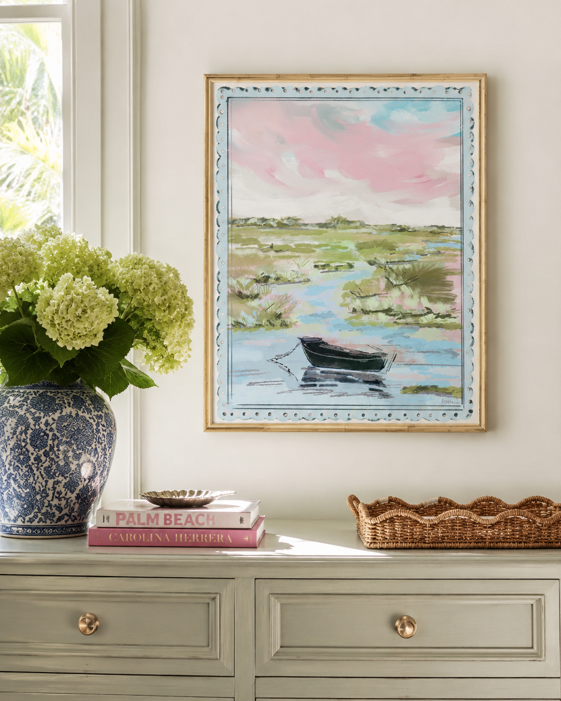 A framed landscape painting of a small boat on a marshy waterway under a pink sky, displayed on a light gray console table in a styled room with a blue patterned vase, books, and a wicker tray.