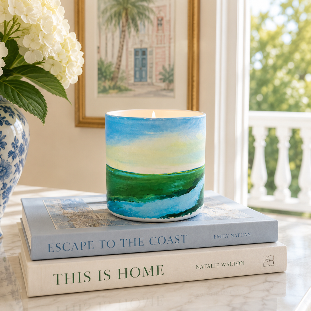 A hand painted cylindrical candle with a coastal landscape design sits on two stacked books on a marble table. The candle features a blue sky, yellow sun, and green sea design.