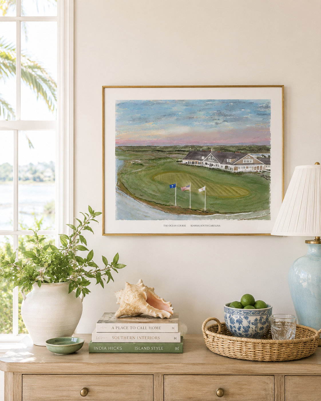 A framed art print depicting a golf course landscape hangs on a beige wall above a wooden console table styled with books, a conch shell, a blue ceramic lamp base, and a bowl of limes.