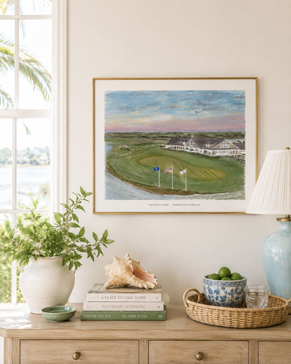A framed art print depicting a golf course landscape hangs on a beige wall above a wooden console table styled with books, a conch shell, a blue ceramic lamp base, and a bowl of limes.