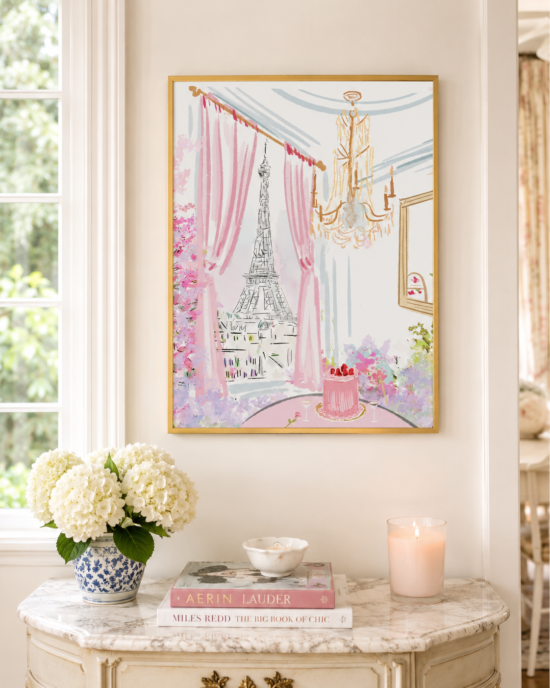A framed watercolor art print depicting a Parisian interior with the Eiffel Tower, pink curtains, and a chandelier, displayed on a cream console table with flowers, books, and a candle in a bright room.