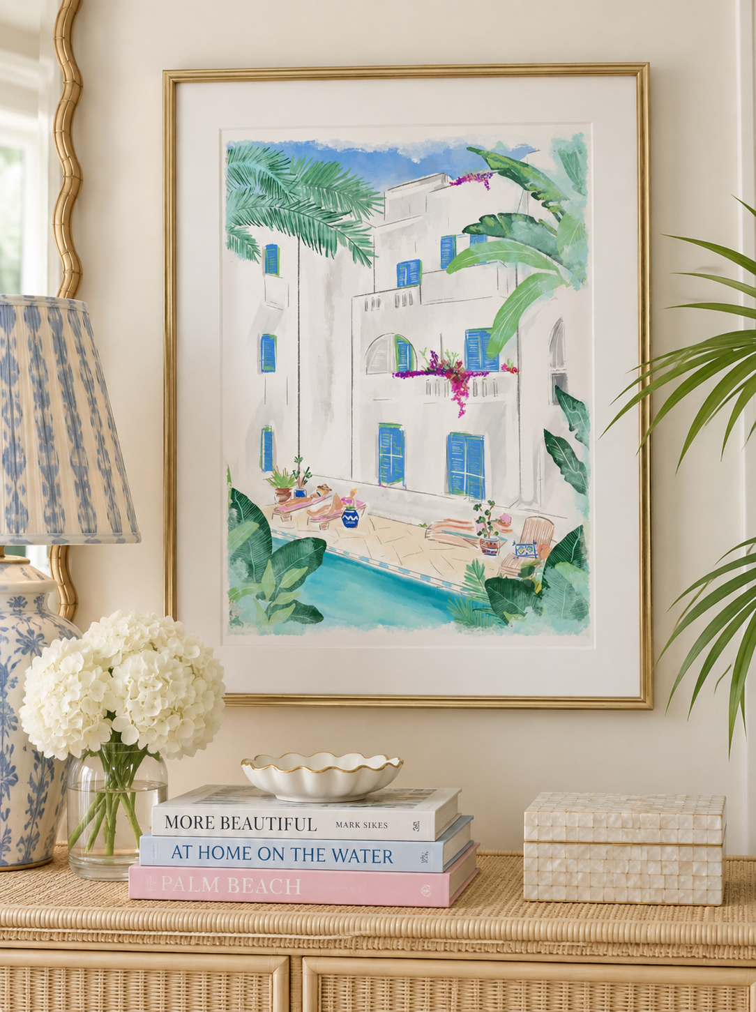 A framed watercolor art print depicting a Mediterranean-style white villa with blue shutters, palm trees, and a pool, displayed on a console table in a styled room setting.