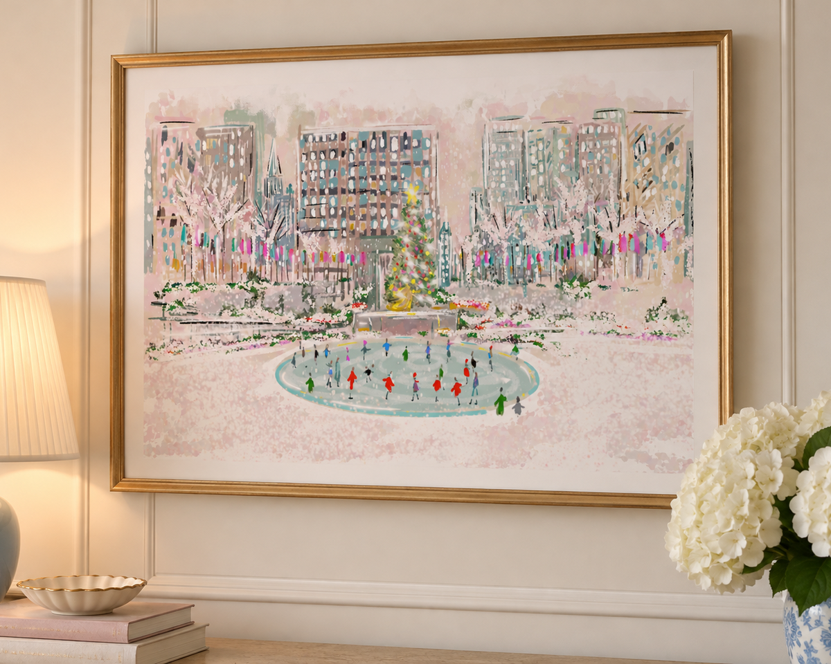 A framed watercolor painting of a winter city scene with a central ice rink, hung on a cream paneled wall above a console table with a lamp, books, and white flowers.