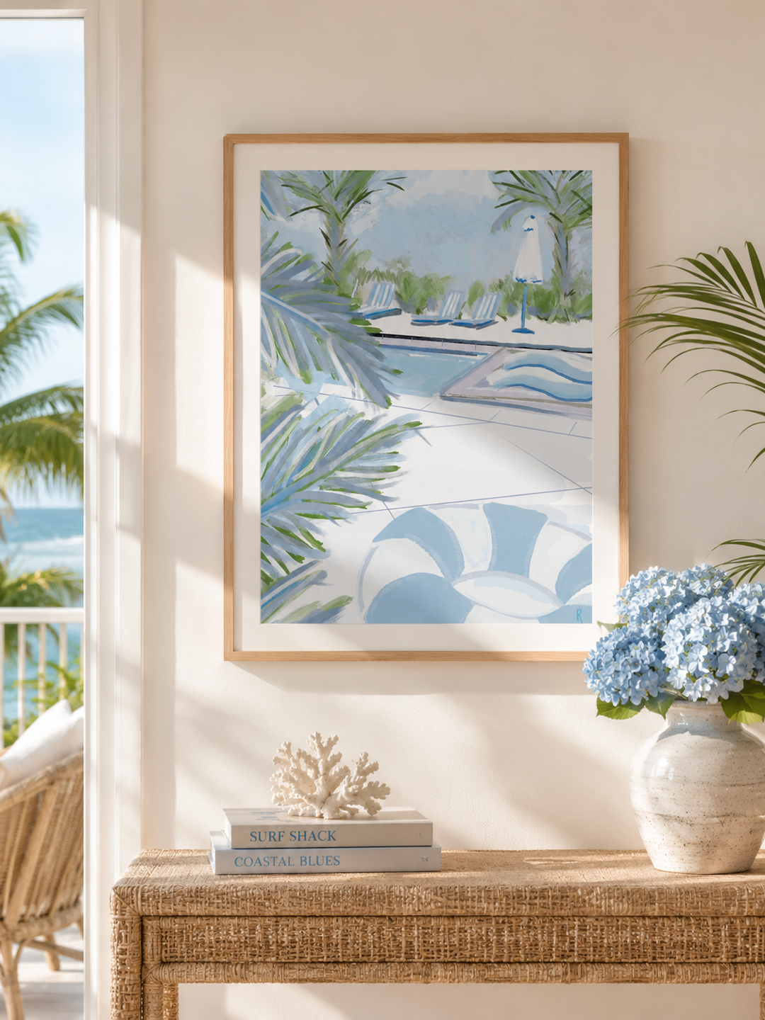 A framed art print featuring a tropical poolside scene with palm leaves and blue lounge chairs, displayed on a beige wall above a woven console table with books, a coral decor piece, and a vase of blue hydrangeas.