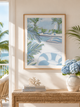 A framed art print featuring a tropical poolside scene with palm leaves and blue lounge chairs, displayed on a beige wall above a woven console table with books, a coral decor piece, and a vase of blue hydrangeas.