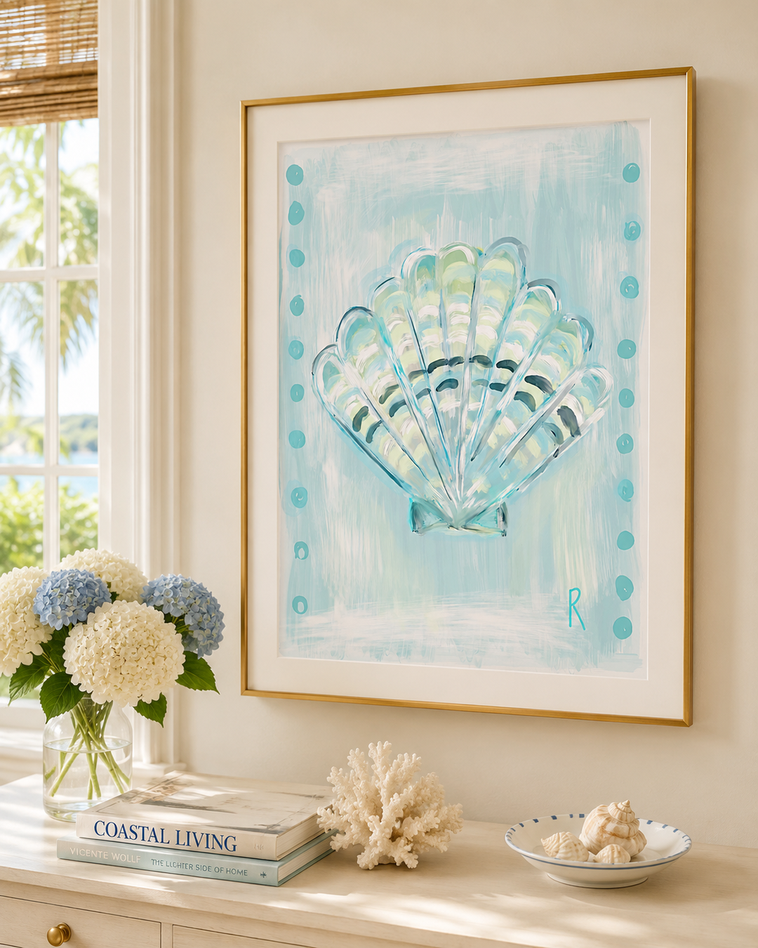 A framed art print featuring a painted seashell design in shades of light blue, teal, and white, styled on a console table in a coastal-themed room with flowers, books, and seashells.