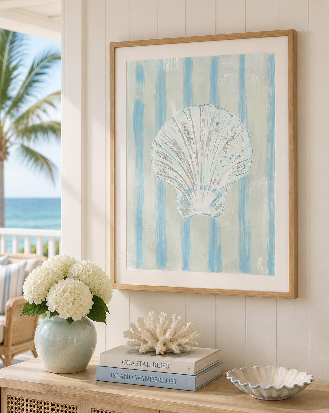Framed wall art featuring a white seashell on light blue vertical stripes, displayed in a light wood frame on a coastal porch with a vase of white flowers, books, and a shell bowl below.