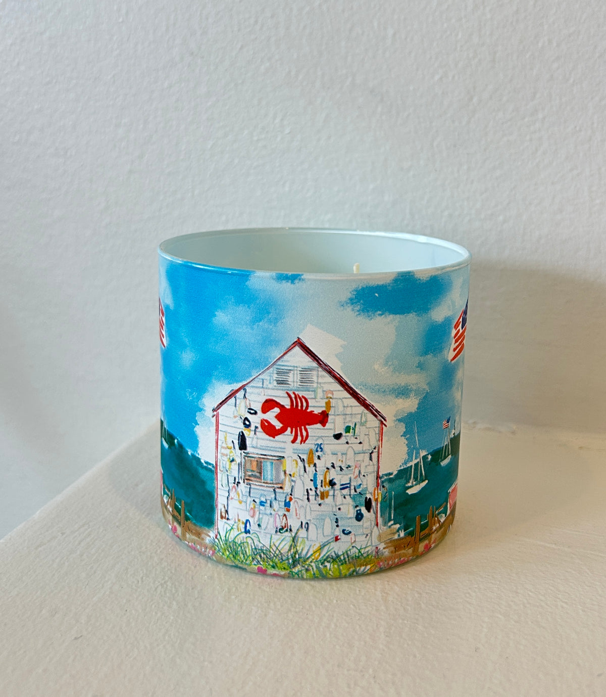 Rebecca Illustrated candle featuring a coastal lobster shack adorned with a red lobster sign, nautical fishing gear, sailboats, and waterfront scenery against a bright blue sky.